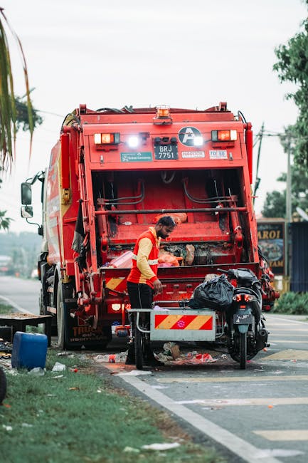A worker in a red and yellow uniform is operating a red waste collection vehicle on a roadside, likely involved in private rubbish removal or on-site clearance. The rear of the truck features an open 
