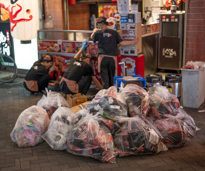 A large, weathered, off-white fabric rubbish bag, visibly worn and stained, is placed on a narrow, uneven urban street pavement near a metal-barred window and a graffiti-marked concrete wall. The bag 