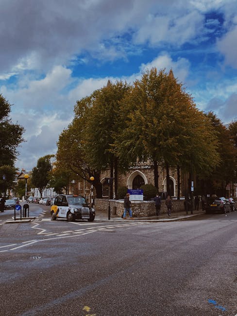 A view of a small church or historic building with a rounded arch entrance, partially obscured by two large trees with dense, autumn-colored foliage. Several parked cars are positioned along the stree